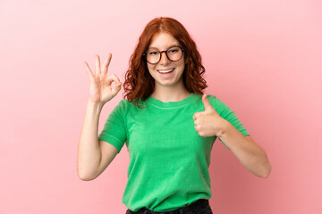 Fototapeta premium Teenager redhead girl over isolated pink background showing ok sign and thumb up gesture