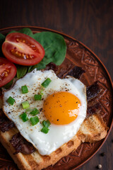 Close-up view of american breakfast consisting of fried eggs with yellow yolk dressed with scallions and pepper on grilled toasted bread with bacon, tomatoes and spinach served on plate. Vertical