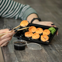 Woman eating hot baked sushi rolls with chopsticks from black plastic container. Takeaway Japanese food concept. Close up shot. Soft focus.