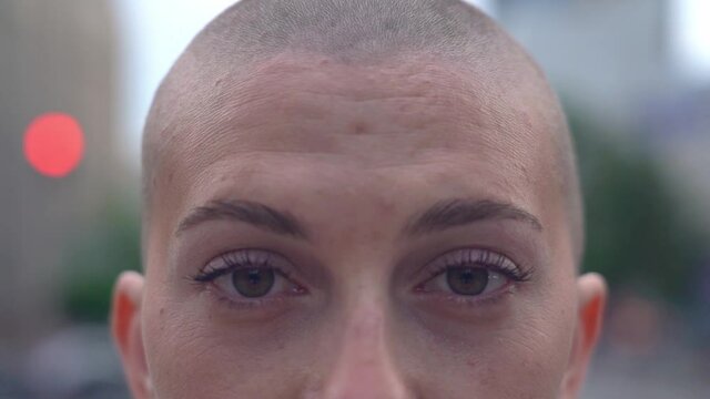 Pensive bald woman looking at camera standing on the street, selective focus. Close up of female eyes blinking