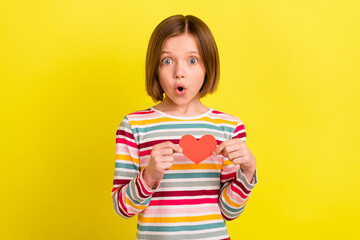 Photo of amazed shocked small girl hold hands small red paper heart shape isolated on yellow color background - obrazy, fototapety, plakaty