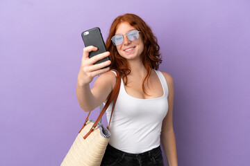 Teenager redhead girl holding a beach bag isolated on purple background making a selfie