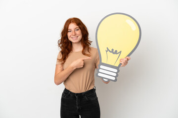 Teenager redhead girl isolated on white background holding a bulb icon and pointing it