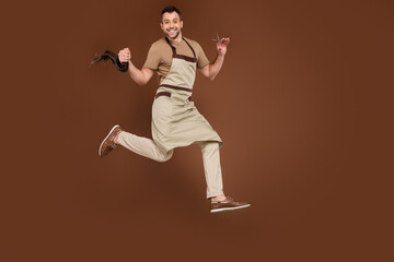 Fototapeta na wymiar Photo of cute funky young gentleman wear beige apron cutting hair tail jumping isolated brown color background