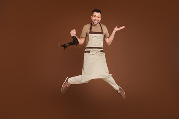 Fototapeta na wymiar Photo of sweet funny young guy dressed beige uniform jumping cutting ponytail isolated brown color background