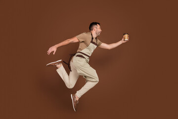 Fototapeta na wymiar Full length body size man wearing apron jumping up giving coffee cup to client isolated brown color background