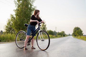 Obraz premium Young woman standing alone on road with bicycle at countryside. Summer activity, healthy lifestyle, workout, sport, fitness. Alone person in nature. Riding bicycle. Having fun outdoors.