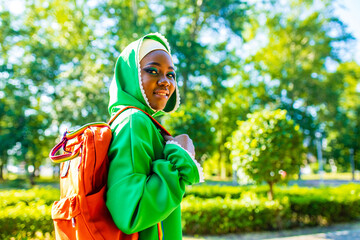 modern afro woman in green hijab with bright make up and piercing nose holg orabge backpack in park