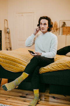 Ung man med h&ouml;rlurar anv&auml;nder smartphone sittande p&aring; s&auml;ng hemma