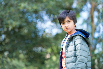 Portrait of Young Indian boy wearing puffer jacket with trees in background.