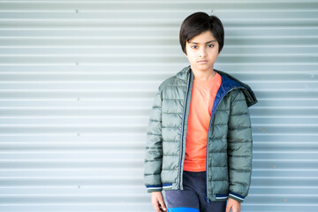 Portrait of young boy looking at camera. Good looking Indian child wearing green puffer jacket standing against corrugated wall.