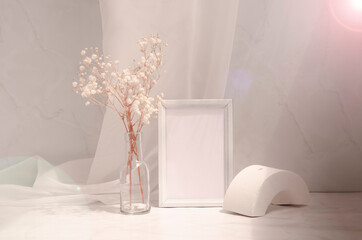Trendy interior still life with sunbeam with glare. Geometric white shape, frame with paper sheet, gypsophila flowers in the glass bottle. Pastel colors background, minimalist design mock up.	