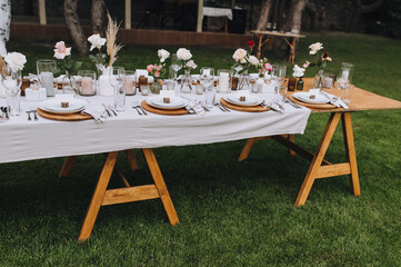 Wooden decorated with flowers, reeds, empty dishes, plates, glasses with a white tablecloth stands on the green grass in the park. Wedding banquet.