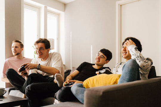 Leende unga v&auml;nner sitter i soffa och spelar tv spel hemma