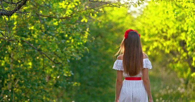 Young woman in a white dress and with long hair is walking in a summer park, the sun's rays illuminate the foliage of the trees. Romantic walk in the summer, beautiful woman, back view. 4k, ProRes