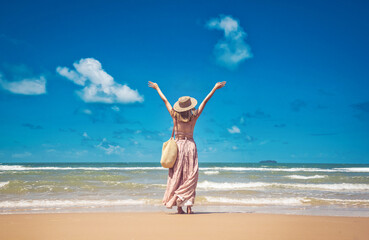 Happy traveller woman in sunhat and dress standing with her arms raised looking tropical beach on summer day