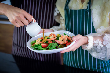 Asian couples in aprons are pouring salad dressing Into a shrimp salad that just helped in the kitchen For eating as a dinner during the holidays