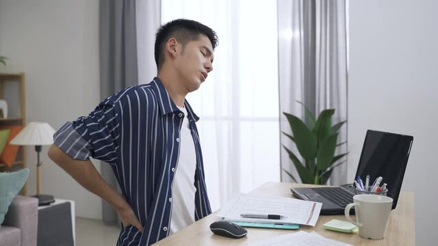 slow motion asian man suffering lower back pain and neck stiffness is taking a break by pounding with fist and rotating head while working from home.