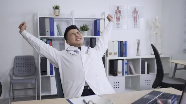 exhausted asian physician putting down his pen on desk is lifting arms to stretch body and exercise stiff neck with a groan while working in the hospital office