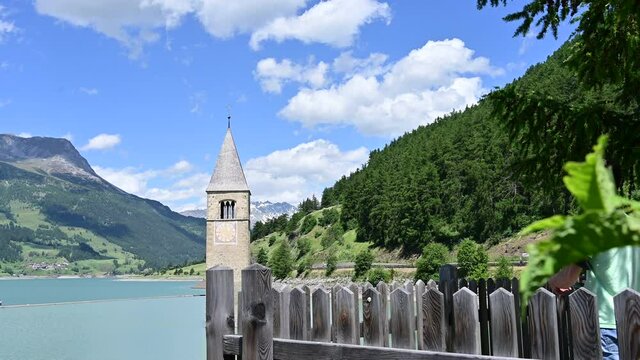 Curon, Italy, June 2021. Footage in beautiful summer day of the old bell tower of Curon (Graun) emerging from the waters of Lake Resia. Two tourists, a woman and a boy, photograph him.