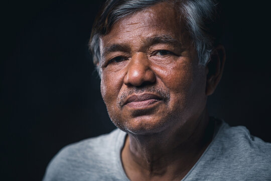 Close up, portrait of Asian elderly man sitting alone looking at camera on dark background, wrinkled skin, gray hair, 60+ years old.