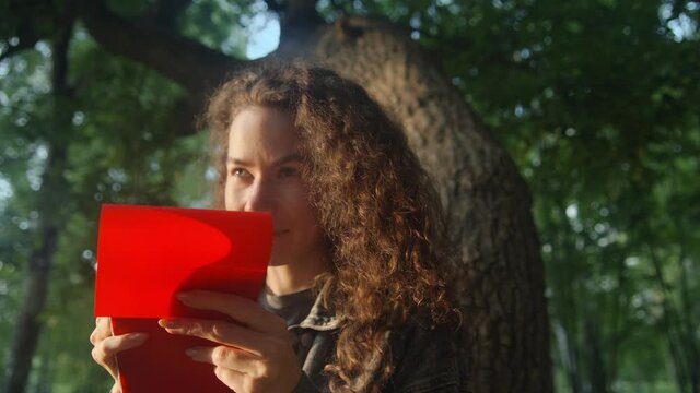 Curly young girl in the park in the summer draws or writes in a red notebook. Painting training in nature. Working with a pencil. 