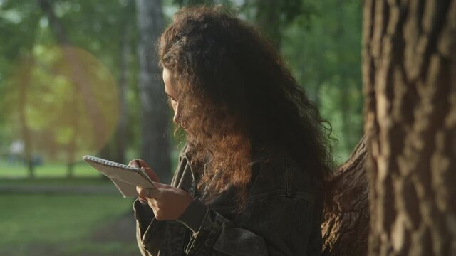 Curly young girl in the park in the summer draws or writes in a red notebook. Painting training in nature. Working with a pencil. 