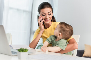 Smiling mother talking on smartphone near toddler son with calculator