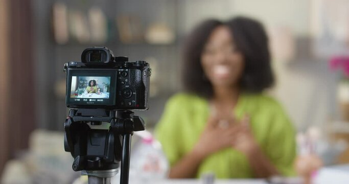 Close up shot of camera recording video of joyful African American young pretty female blogger sitting at home talking to camera about beauty products. Fashion vlog, recording beauty webinar