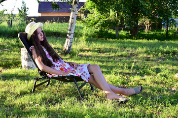 pretty girl with long wavy hair in hat is sitting on chaise longue against  background of rural...