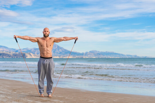 Focused bearded man working out with elastic bands on coast