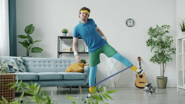 Slow motion of funny sportsman training with resistance band feeling tired and lazy exercising alone at home. Sports activities and humor concept.