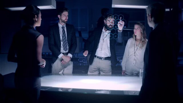 Businessman in suit talking to business people colleagues or partners standing around futuristic table with light showing an architecture project powered by a holographic modern projection technology.