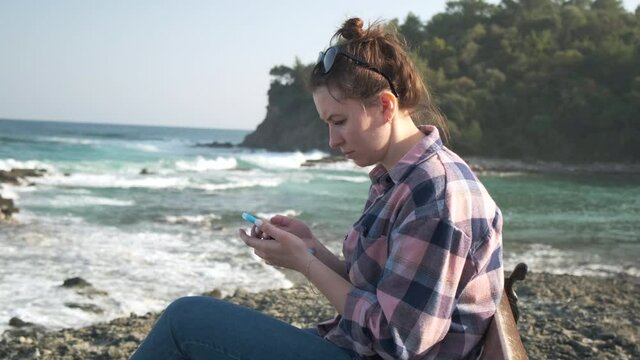 Girl Sits on Rocky Beach. Sea Waves Splashing in Gulf. Holds Phone in Hands. Reads the News. Overwrites. Leads Travel Blog. Post New Vacation Photos. Bragging about Traveling Abroad. Sea Surface
