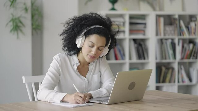 Portrait of African American woman talking on video conference call using laptop and headphones taking notes on notepad. Brunette sits at table in home office. Close up. Slow motion ready 59.97fps.