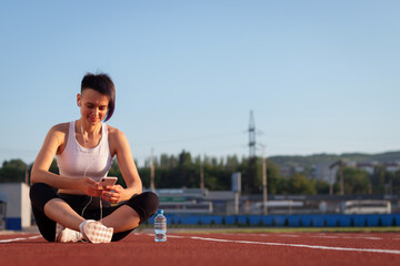 Fototapeta na wymiar A cheerful young woman in sportswear relaxing after a workout or run, sitting on a stadium track, taking selfies, listening to music on headphones on her smartphone, taking a break during a workout