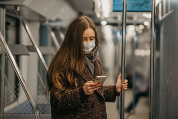 A woman in a medical face mask to avoid the spread of coronavirus is standing and using a smartphone in a modern subway car. A girl in a surgical mask is scrolling news on her cellphone on a train.