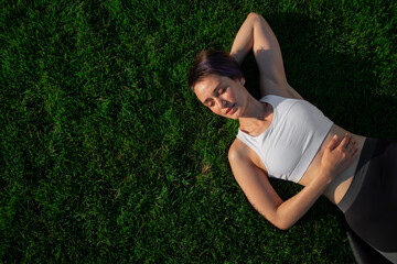 A beautiful, cheerful, cheerful woman in a white T-shirt is lying on the green lawn in the park and smiling. Happy woman relaxing on the grass during sunny summer day. Top view. large format banner