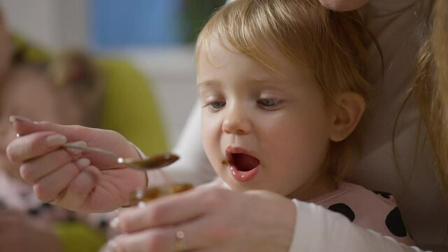 Close-up portrait of blond beautiful baby girl with grey eyes eating multivitamin nutritious food from spoon. Unrecognizable young mother feeding cute daughter as twin eating at background