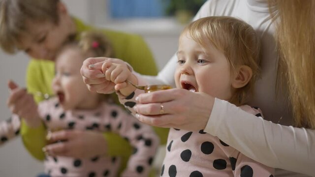 Portrait of cute little girl eating baby food at home as twin sister enjoying lunch at background. Unrecognizable mother and blurred grandmother feeding children indoors