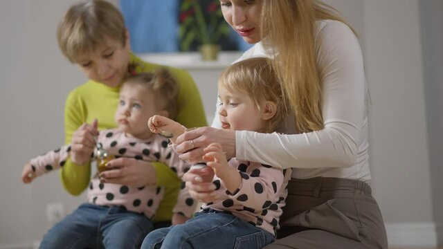 Side view of unrecognizable mother giving spoon for infant eating as grandmother feeding twin sister at background. Siblings eating baby food in the evening at home