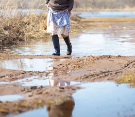 a woman in a long dress and rubber boots crosses mud and puddles after a river flood, a spring day