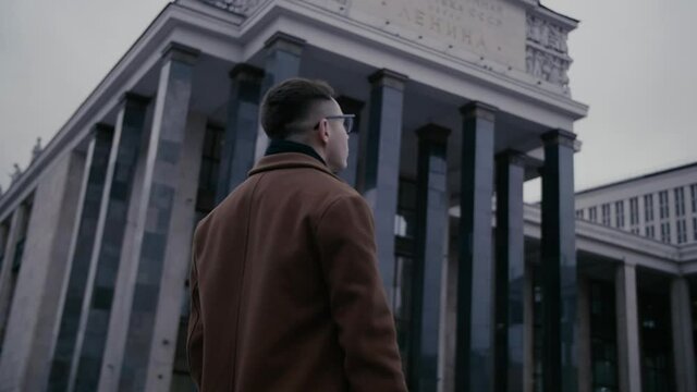 Young businessman looking at Russian State Library. He's wearing a brown long coat. Back view of businessman in coat outdoors on street. Stylish male entrepreneur in warm clothes going to work outside