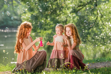 Three red-haired sisters in long linen dresses blow bubbles in the park on sunny summer day.