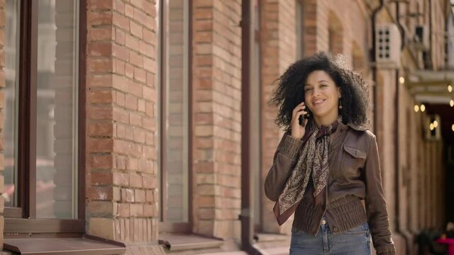 Portrait of young African American woman talking on smartphone. Brunette with curly hair in leather jacket walks down street against backdrop of beautiful brick building. Slow motion ready 59.97fps.