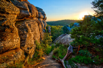 dirt path down through a canyon beside a sun colored cliff face reaching to the sky, small cedar, grass, and dead wood on the right, sun setting on distant mountain ridge, Shawnee Nat. For. Illinois