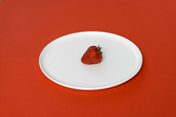 One juicy red strawberry lies on a white plate on a red background (927)