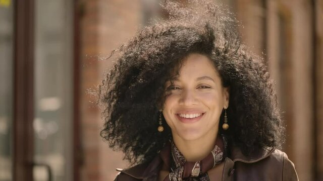 Portrait of stylish young African American woman flirtatious smiling and winking. Brunette in brown leather jacket posing on street against blurred brick building. Close up. Slow motion ready 59.97fps
