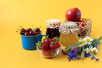 The concept of collecting and processing summer vitamin berries. Jam jars, strawberries, cherries, apples, a bouquet of wild flowers on a yellow background, side view, space for text