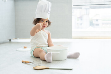 Portrait of little Caucasian adorable kid girl wear chef hat holding wooden spoon and tasting food from pot with white t-shirt mess up with ketchup. Cute child sitting on floor at kitchen at home.